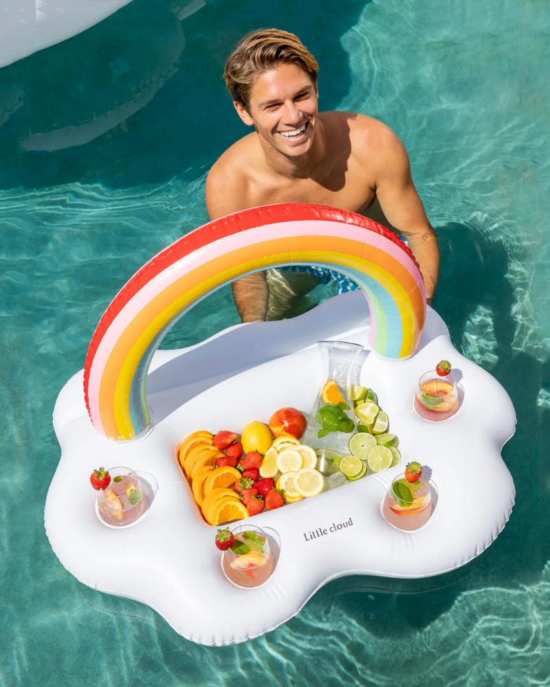 A man standing in a swimming pool holding a Little Cloud rainbow inflatable tray (cloud-shaped pool drink float) filled with citrus fruit slices and drinks, enjoying a summer pool party